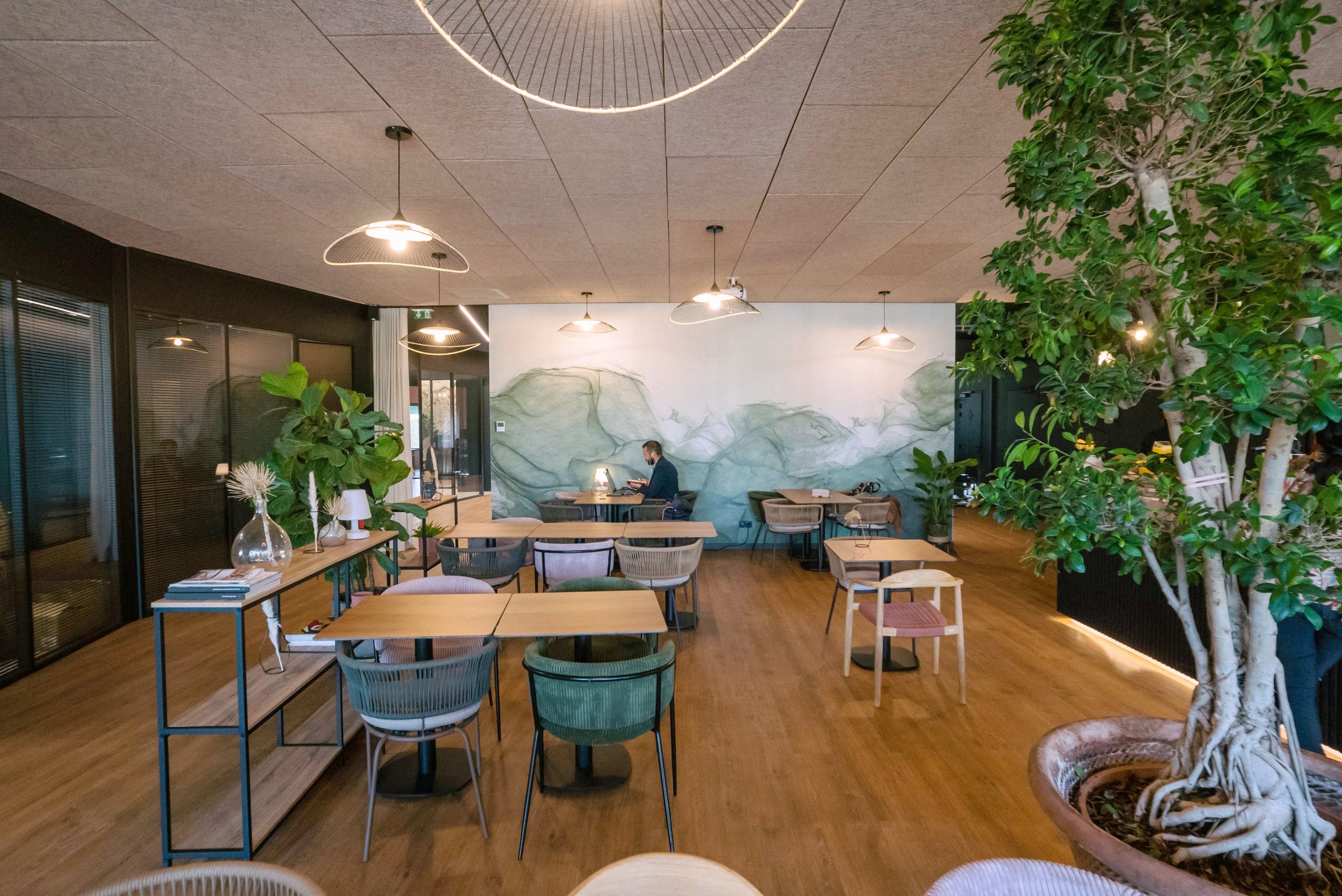 Espace lounge de La Gravade : tables en bois, fauteuils, mur végétal peint et grand ficus, ambiance chaleureuse et vivante